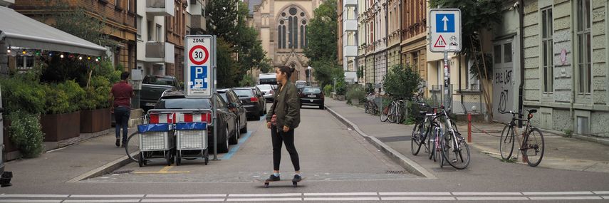 Frau auf Skateboard vor Quartierstrasse mit parkierten Autos und Velos, im Hintergrund ist eine Kirche sichtbar.